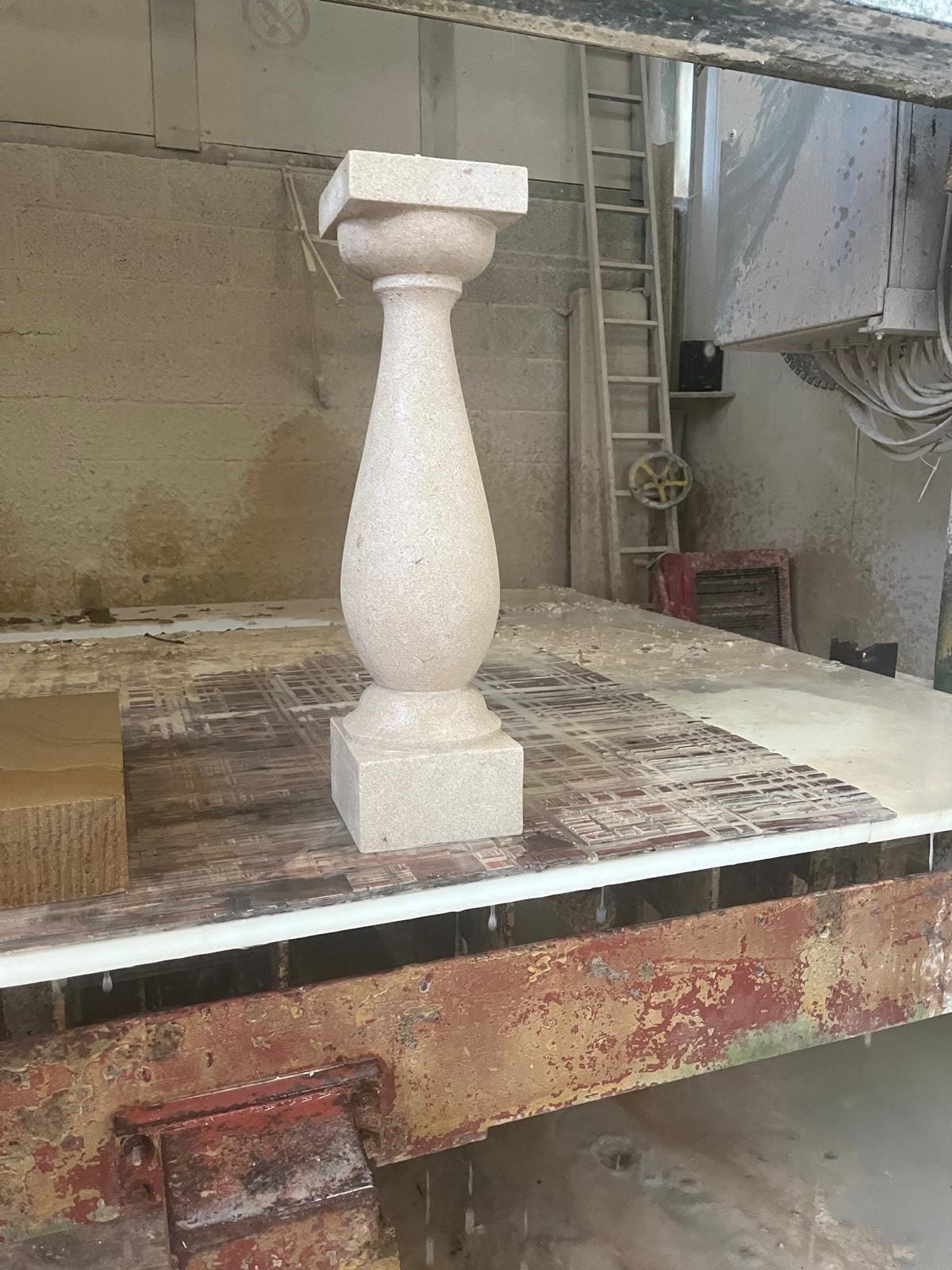 A turned stone baluster with a square base and cap standing on the bed of a workshop saw