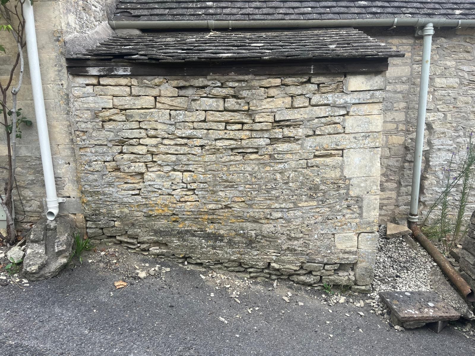 A Cotswold stone elevation before repointing, the joints eroded and open, the face darkened with algae and loose mortar fallen at the base