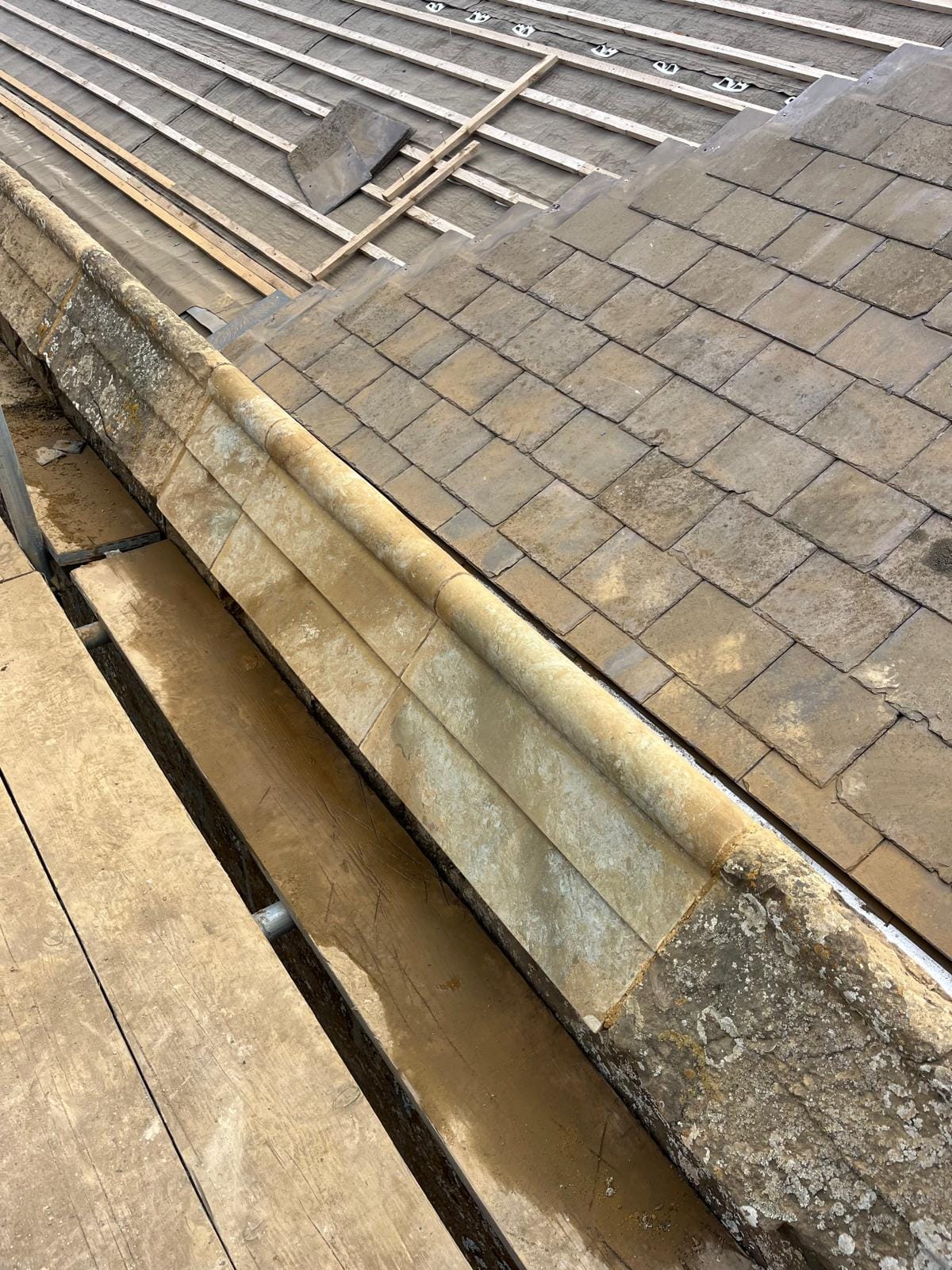 A rounded stone coping running along a roof verge beside newly laid stone slates, seen from the scaffold