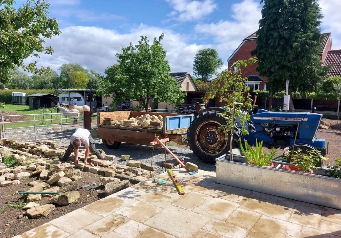 Dry stone wall under construction beside a garden, with a trailer of Cotswold stone and a tractor alongside and horses in the paddock beyond