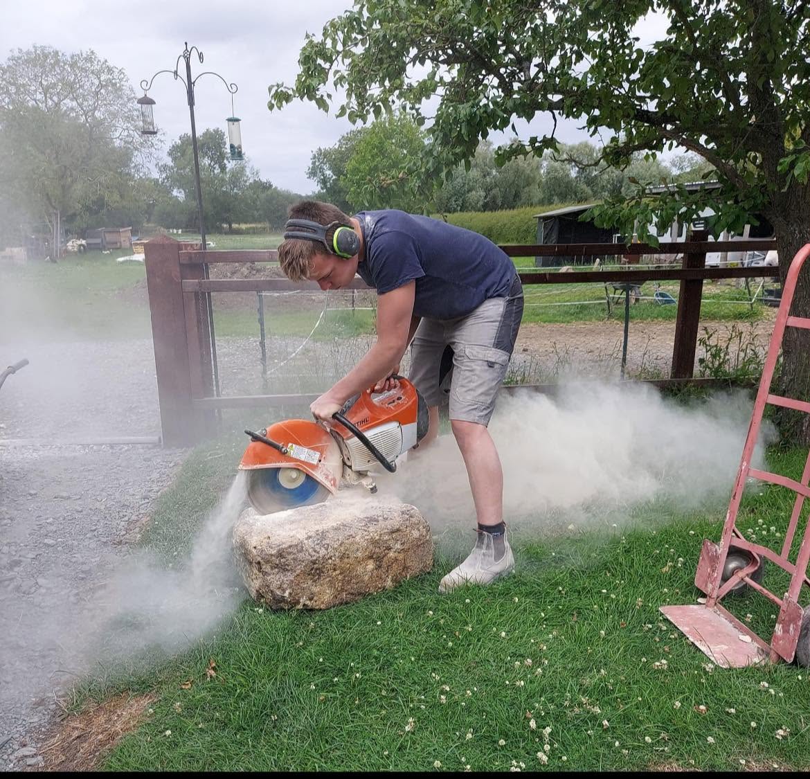 Charlie in ear defenders cutting a large block of limestone with a petrol disc saw, dust billowing across the grass