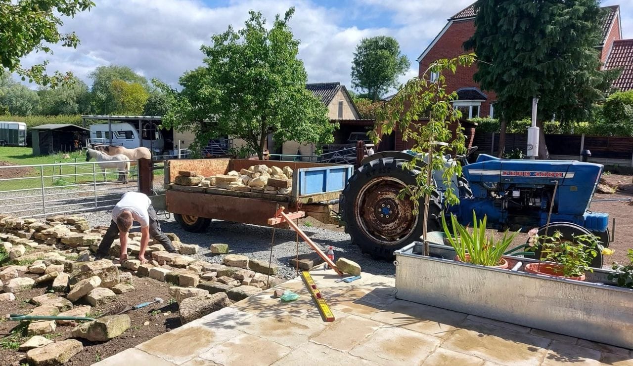 Charlie building a dry stone wall in a garden, with stone graded out along the line of the wall and a tractor and trailer of stone behind